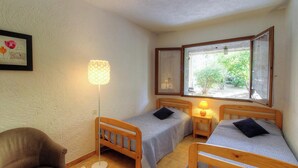 Una habitación pequeña y acogedora con dos camas individuales, una mesita de noche con lámpara, una silla y un ventanal con vistas a la vegetación.