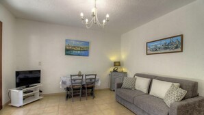 Una sala de estar con televisor, mesa de comedor, sillas, sofá, candelabro y dos cuadros en las paredes.