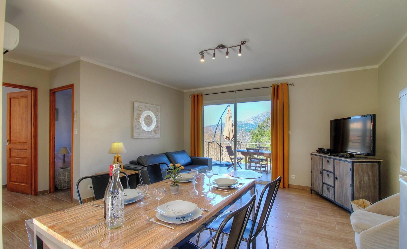 Un coin repas avec une table dressée pour quatre personnes, un canapé, un téléviseur posé sur un meuble et une vue dégagée sur l’extérieur grâce à une grande fenêtre.