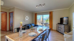 Ein Essbereich mit einem Tisch für vier Personen, einer Couch, einem Fernseher auf einem Schrank und Blick durch ein großes Fenster.