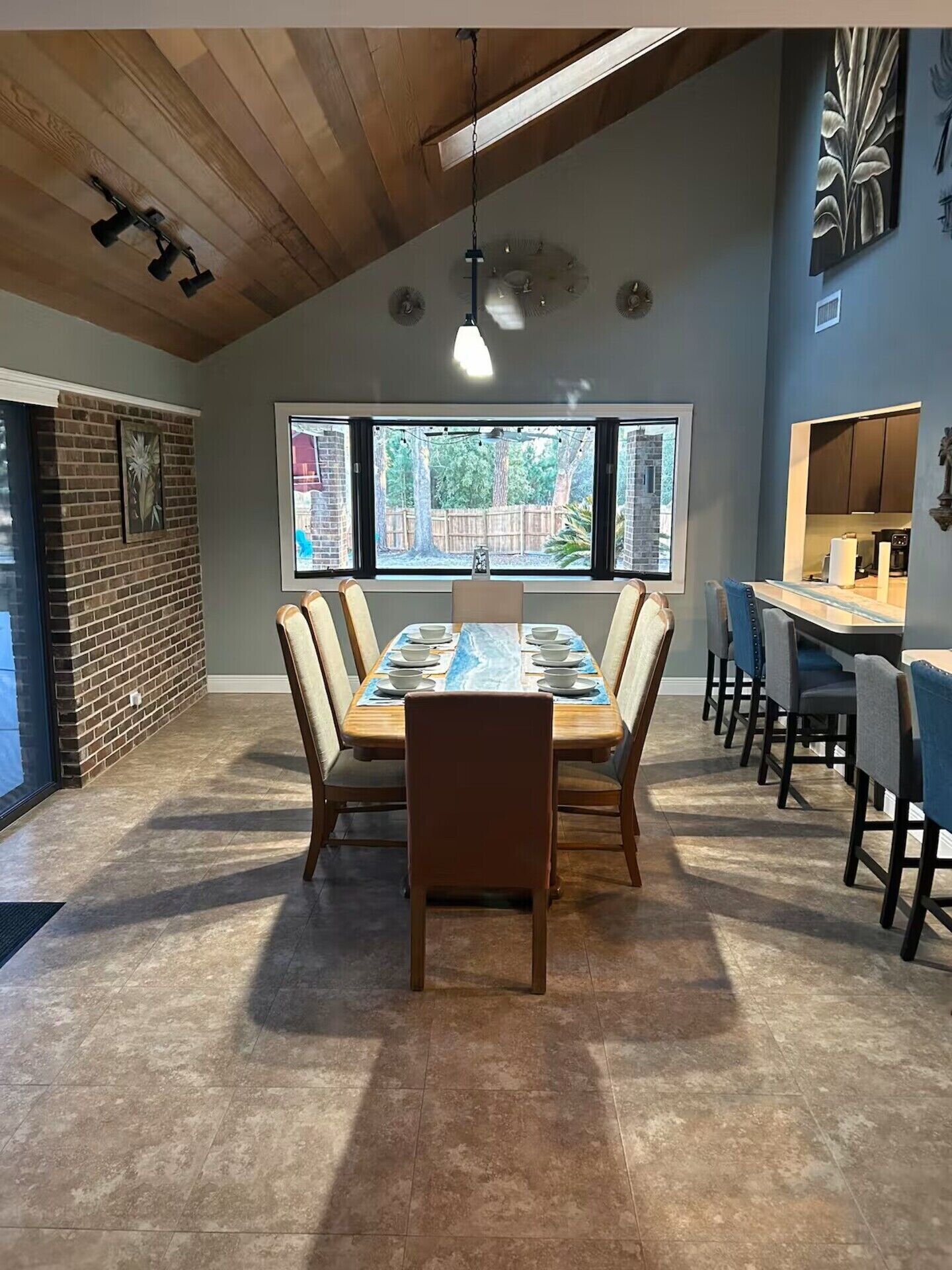 A dining area with a long table, chairs, a hanging light fixture, a brick wall, and a window with a view of trees.