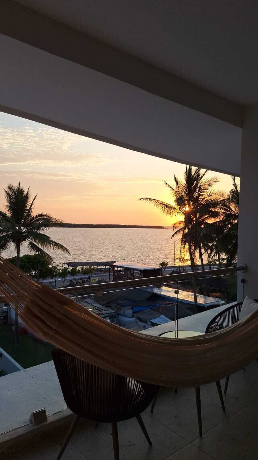 A hammock on a balcony overlooking a pool and palm trees at sunset.