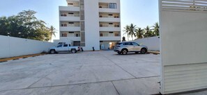 Un área de estacionamiento con una camioneta pickup blanca y una SUV plateada, un edificio alto con varios balcones, y palmeras al fondo.
