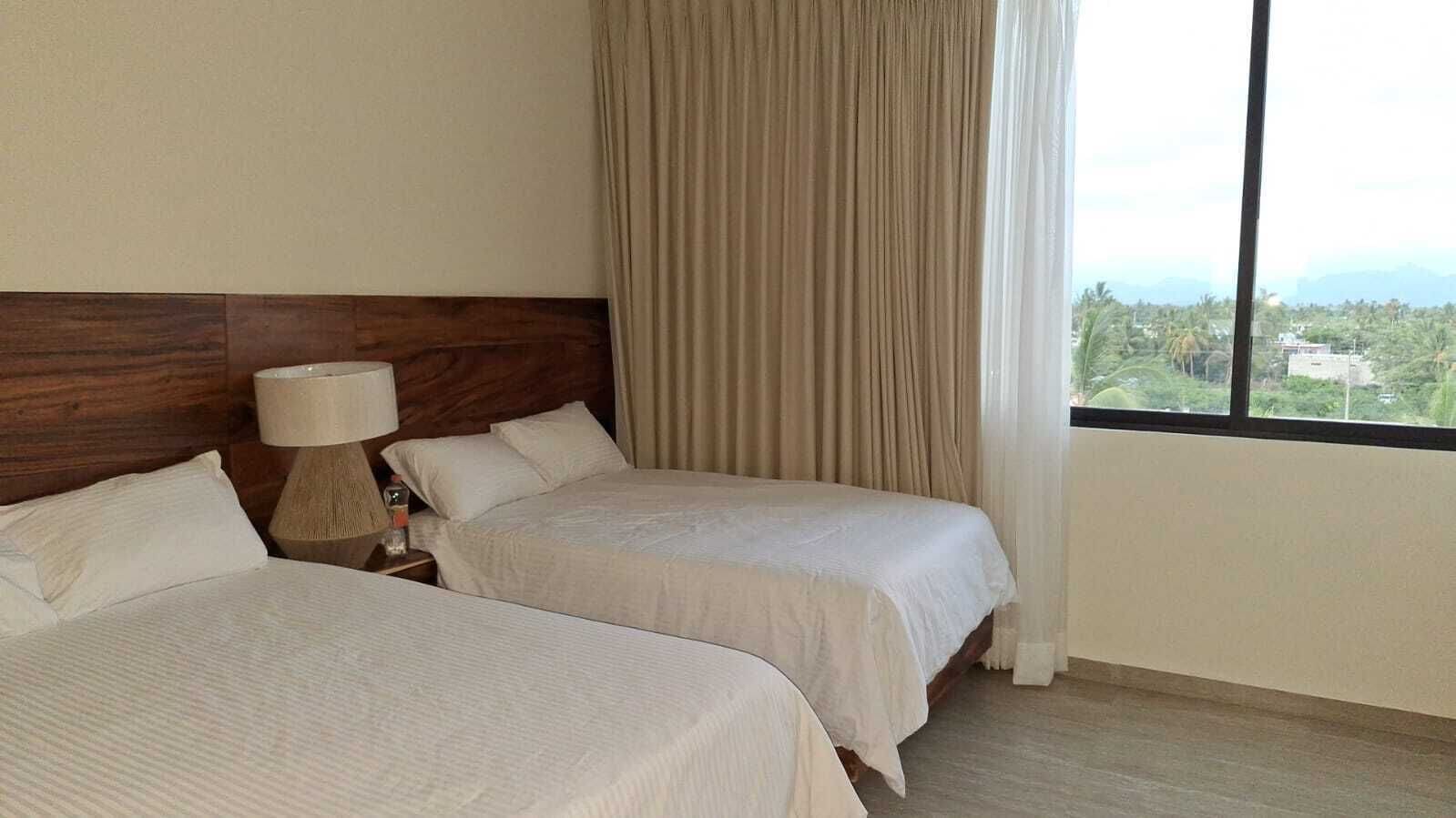 Habitación de hotel con dos camas, cabecero de madera, lámpara de noche y ventana con vista a follaje.
