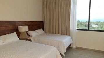 Habitación de hotel con dos camas, cabecero de madera, lámpara de noche y ventana con vista a follaje.