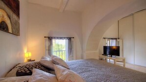 A bedroom with a bed, a television on a stand, a window with curtains, and a painting on the wall.