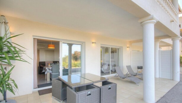 Un patio esterno moderno con tavolo in vetro, sedie in vimini e una porta scorrevole che dà su una cucina.
