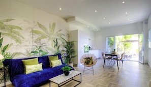 Ein Wohnzimmer mit einem blauen Sofa, einem Essbereich mit Tisch und Stühlen sowie einer Küche im Hintergrund.