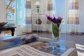 Ein Zimmer mit Glastisch, auf dem eine Zeitung liegt, einer Vase mit violetten Blumen und einem Metallregal mit einer Lampe.