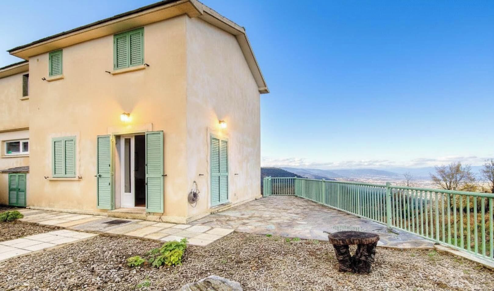 A two-story building with a stone patio, green shutters, and a view of distant mountains.