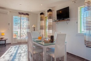 Een eethoek met een glazen tafel, witte stoelen, een aan de muur bevestigde televisie en gestreepte gordijnen.