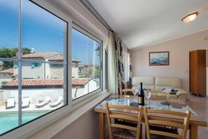 Ein Zimmer mit Glastisch, Stühlen, einer Couch, einem Gemälde und Blick auf einen Swimmingpool und Gebäude.
