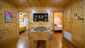 Habitación con piso de madera, una mesa de fútbol de mesa, un estante con objetos decorativos y una cama al fondo.