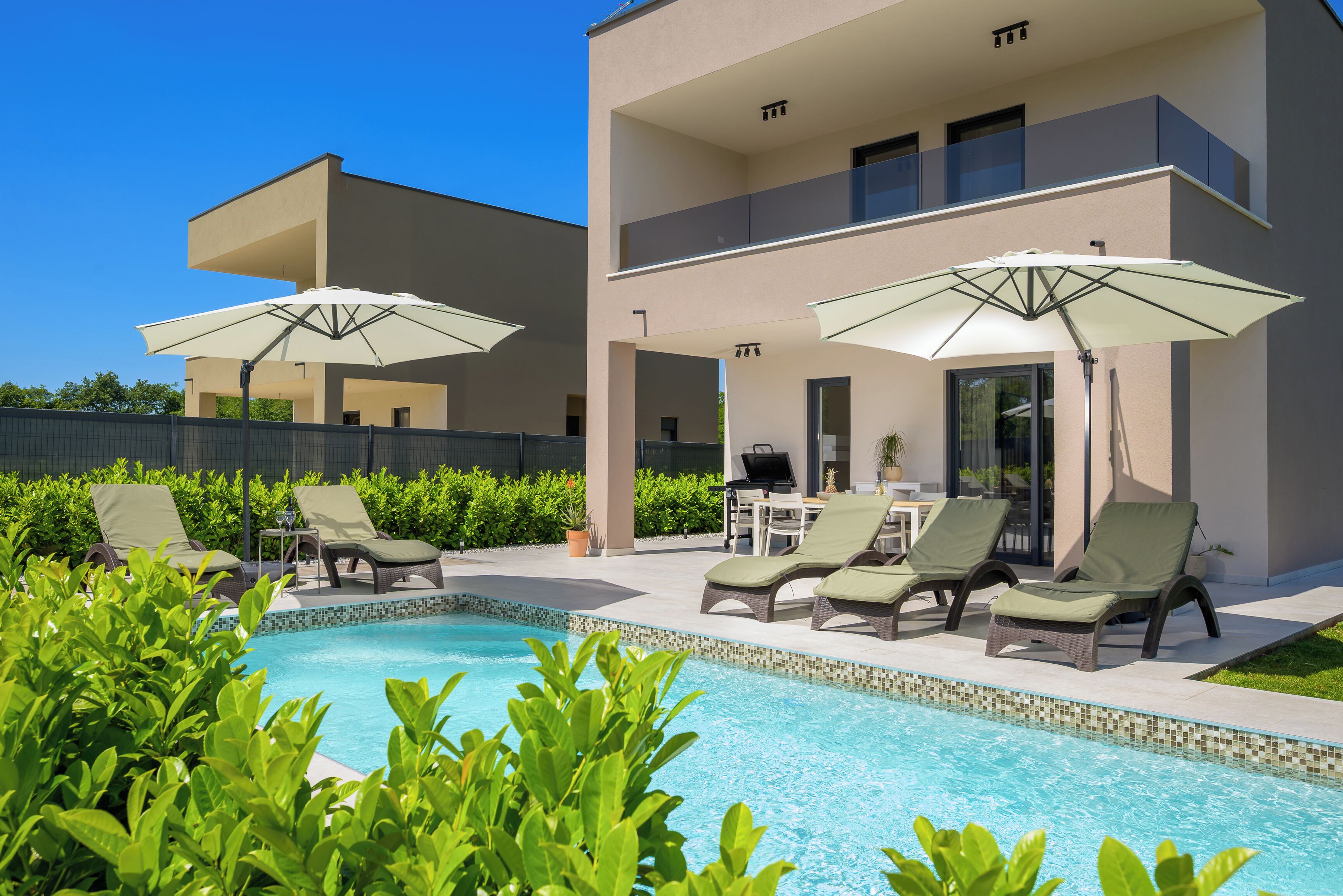 Un espace piscine avec des chaises longues et des parasols, un coin barbecue et un bâtiment à deux étages doté de balcons.
