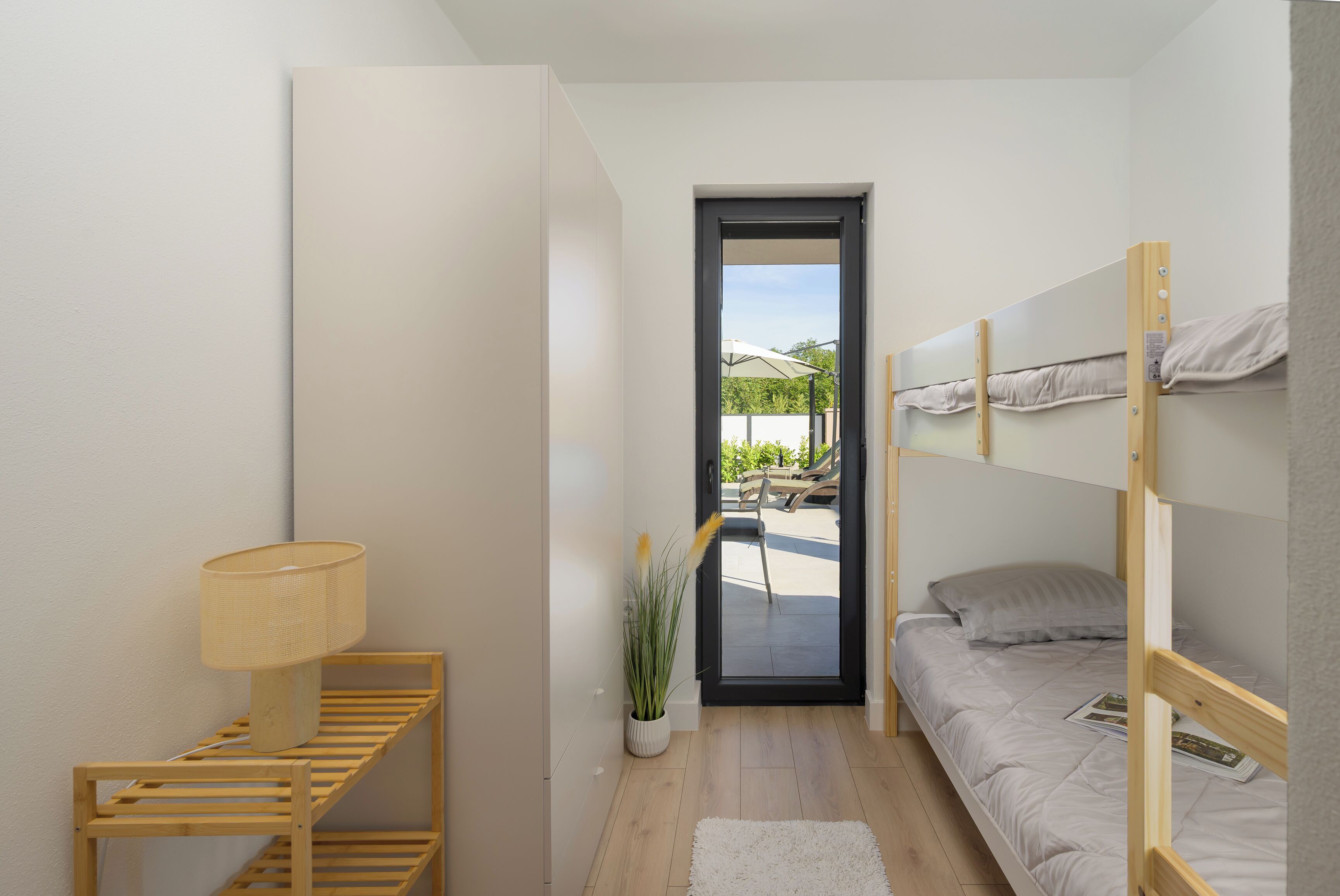 A compact room with a bunk bed, a desk with a lamp, a potted plant, and a view through an open door.