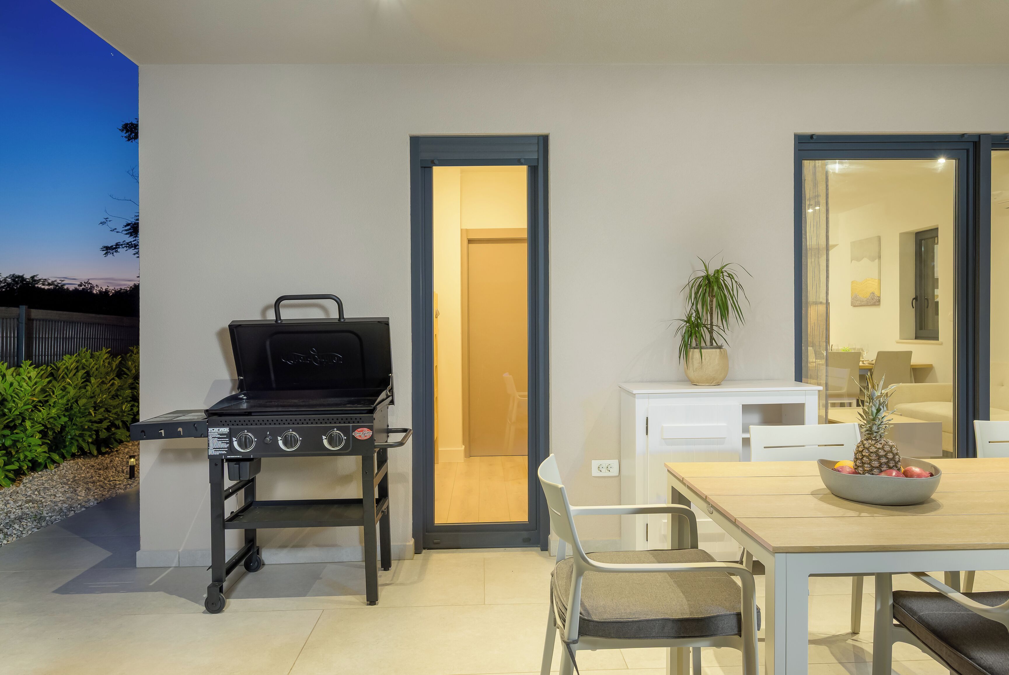 Une terrasse extérieure avec un barbecue, une table à manger avec des chaises, un pot de fleurs et une vue sur la maison.