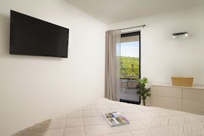 Ett sovrum med en säng, en platt-TV som är monterad på väggen, ett fönster med gardiner, ett litet bord med en växt och en korg på en byrå.