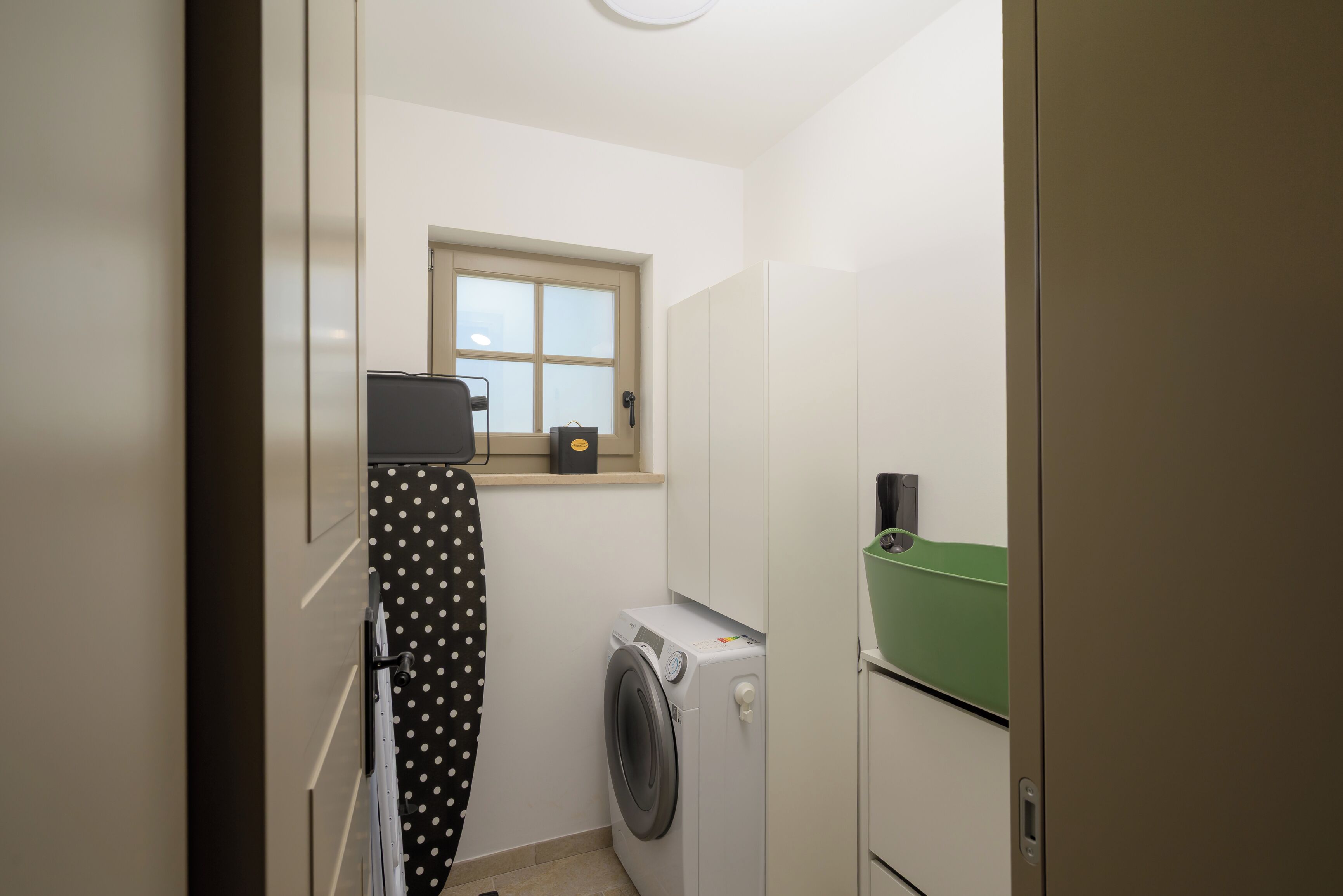 Een kleine wasruimte met een wasmachine, een droger, een klein raam en een groene mand op het aanrecht.