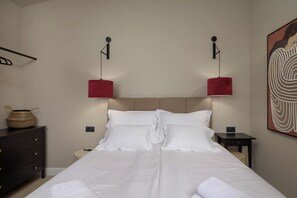 Ein ordentlich bezogenes Bett mit weißen Leinen, eine dunkle Holzkommode mit Schubladen, ein kleiner Nachttisch mit Lampe und ein an der Wand befestigtes Kunstwerk.