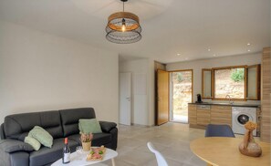 A modern living room with a sofa, coffee table, and dining area. There is a kitchen with a washing machine and a large window. - Les Bergeries de Paomia (CARGESE)