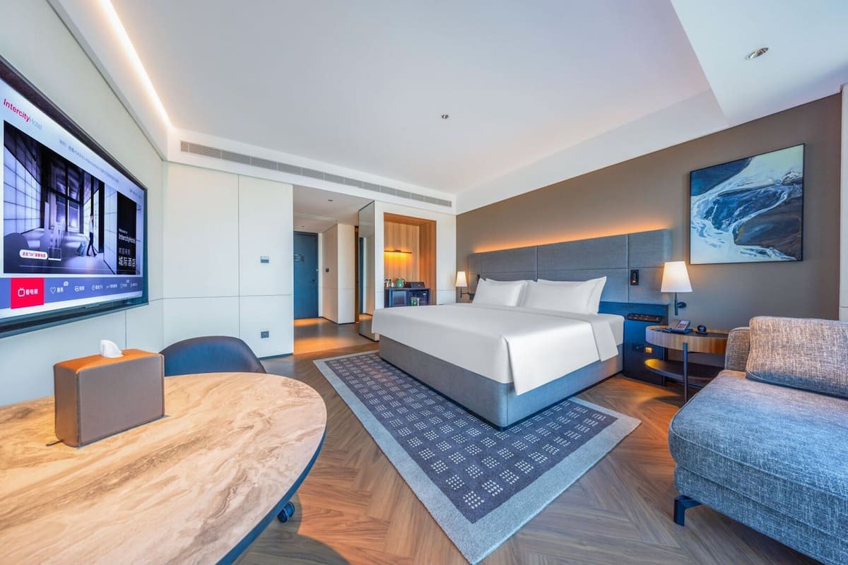 Una habitación de hotel moderna con una cama grande, un escritorio, una silla, un televisor y un cuadro en la pared.