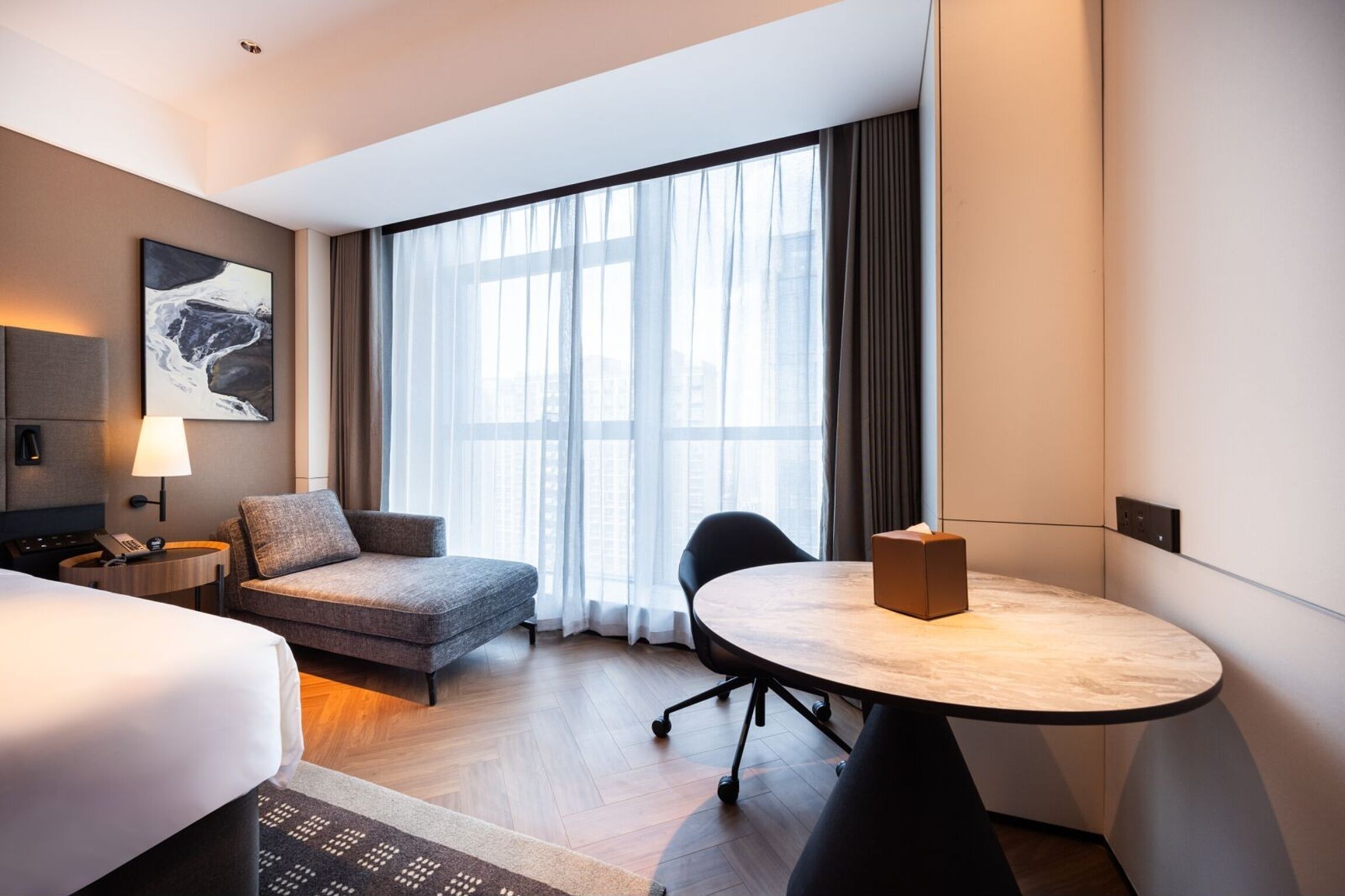 Habitación de hotel con cama, sofá, una mesa redonda, lámpara, un cuadro en la pared y un ventanal con cortinas.