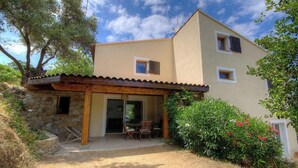 Un bâtiment à deux étages, avec une structure en pierre sur le côté gauche, un patio couvert avec des chaises et un jardin agrémenté d’arbustes et de fleurs.