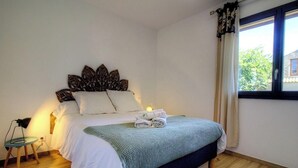 A bedroom with a bed, pillows, a bedside lamp, a window with curtains, and a small table with a chair. - Le grand gîte d'Elodie (TALLONE)