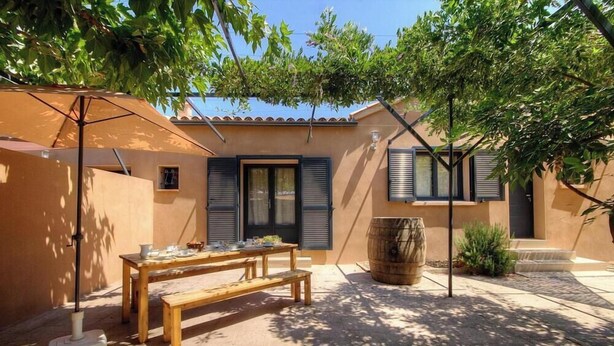 A patio with a wooden table and benches, a barrel, and a pergola with an umbrella. - Le grand gîte d'Elodie (TALLONE)
