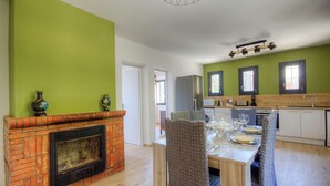 A dining area with a brick fireplace, a table set for four, and a kitchen in the background. - Le grand gîte d'Elodie (TALLONE)