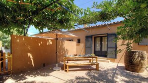 A patio with a wooden table and benches, a wooden barrel, and a house with a tiled roof and shutters. - Le grand gîte d'Elodie (TALLONE)