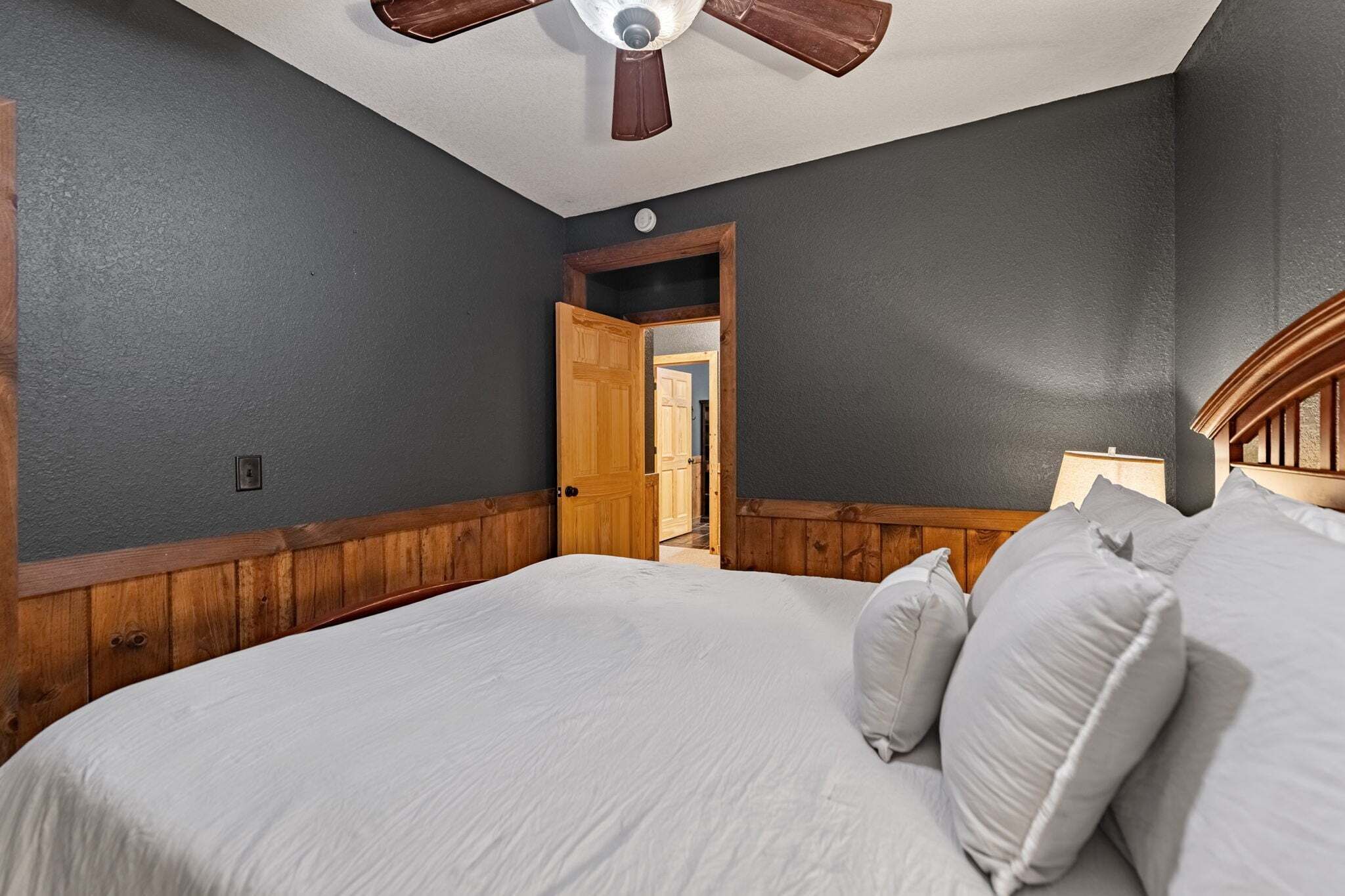 A bedroom with a bed, wooden paneling, a ceiling fan, and a door leading to another room.