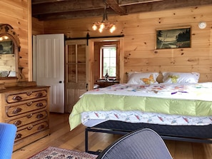 A wooden cabin interior with a bed, dresser, chair, and a painting on the wall.