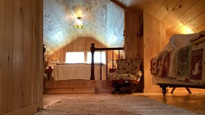 A cozy attic bedroom with a wooden ceiling, a bed with a quilt, a chair, and a large window.