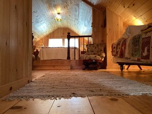 Tunnelmallinen ullakkohuone, jossa on puinen katto, peitolla varustettu sänky, tuoli ja suuri ikkuna.