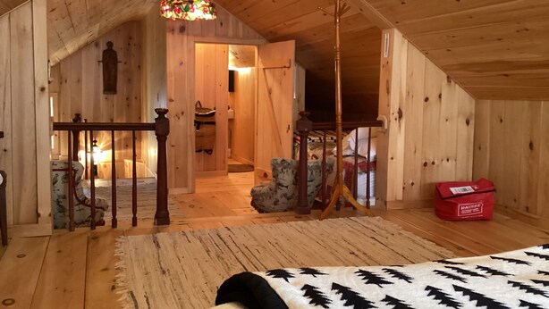 A wooden interior with a sloped ceiling, a staircase, a hanging light fixture, and a bed with a patterned blanket.