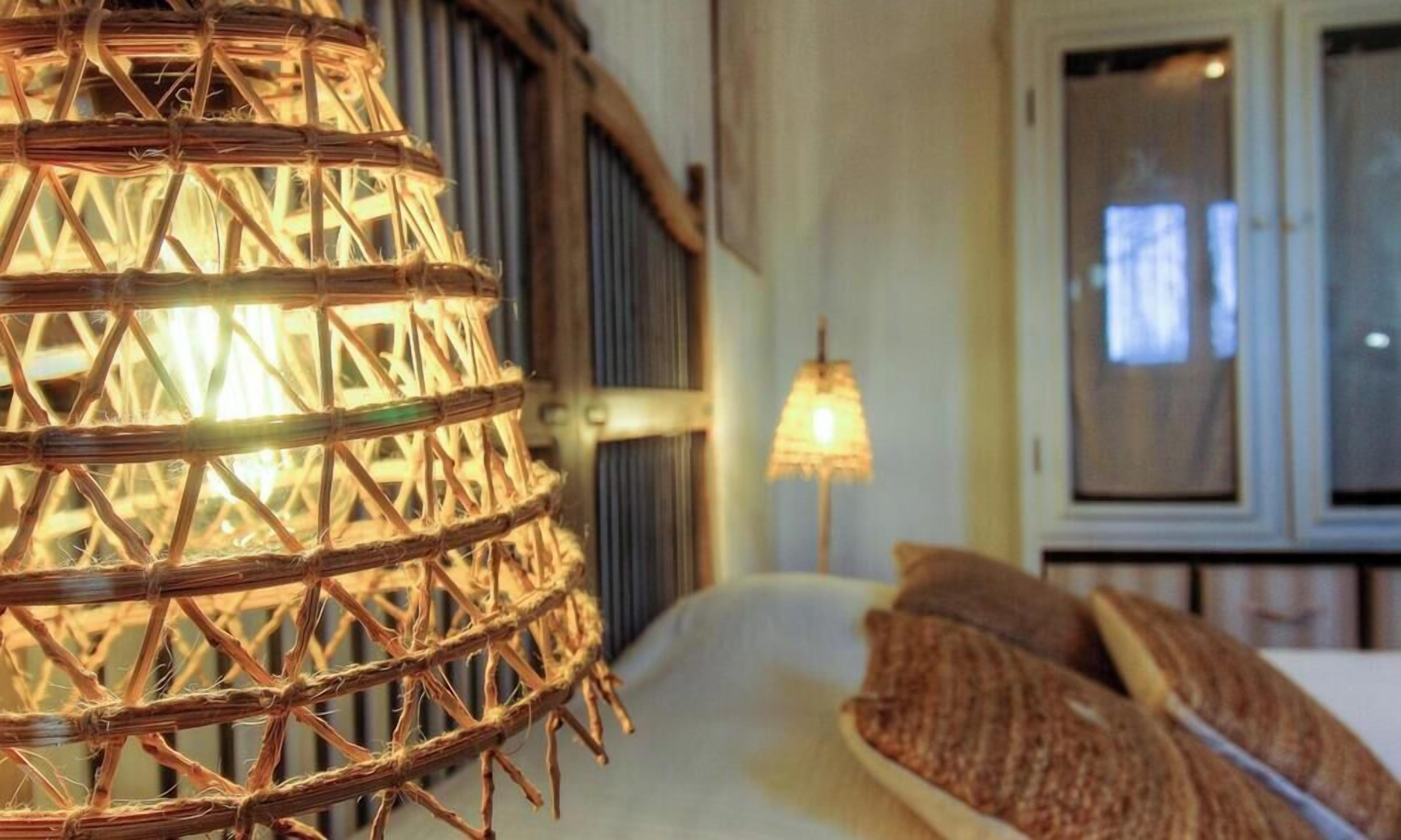 Una lámpara de mimbre, una cama con almohadas y una ventana con persianas.