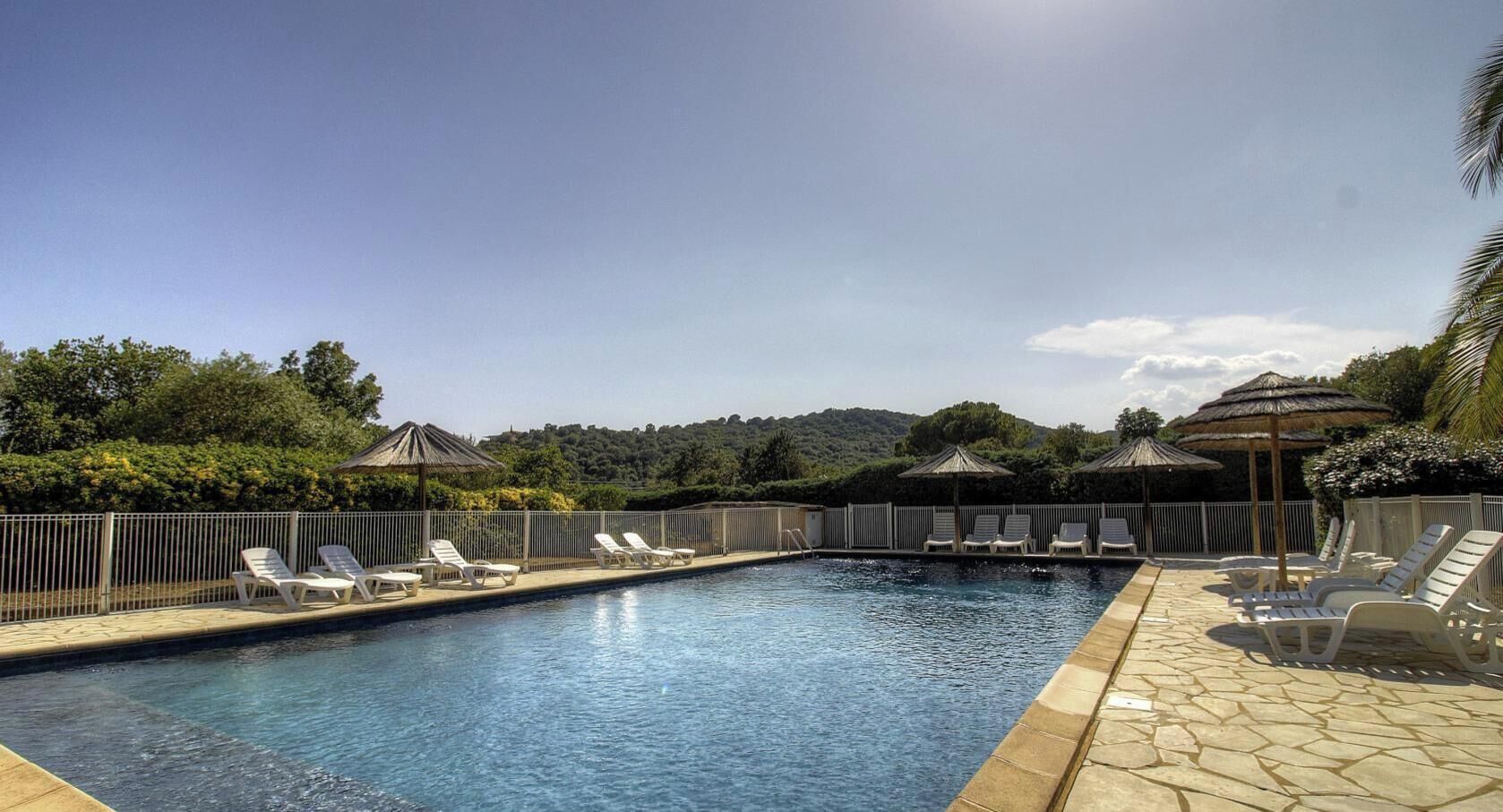 Ein Schwimmbecken mit Liegestühlen und Sonnenschirmen, umgeben von einer Hecke und Grünpflanzen.