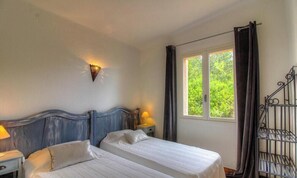 Ein Zimmer mit zwei Betten, einem Nachttisch mit Lampe, einer Wandleuchte, einem Fenster mit Vorhängen und einem Metallgitter.