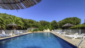 Ein Swimmingpool mit weißen Liegestühlen und Strohdächern, umgeben von viel Grün.
