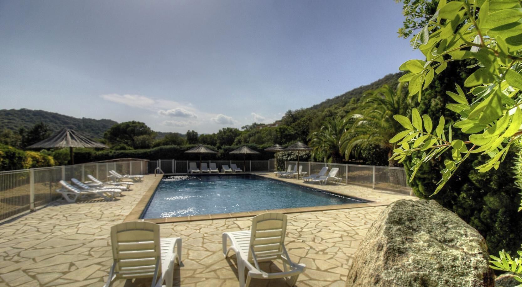 A rectangular pool with a stone deck, surrounded by lounge chairs and umbrellas, set against a backdrop of greenery and hills.