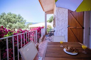 Ein Holztisch mit einer Platte Croissants und einem Glas Orangensaft, ein weißer Stuhl sowie ein Balkon mit Geländer, der auf einen Garten mit rosa Blumen blickt.