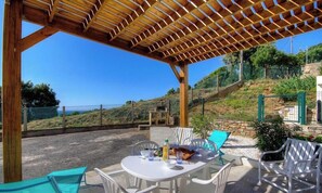 An outdoor patio with a wooden pergola, white chairs, a table with food, and a view of a hillside. - Le 62861 - FARINOLE - Gîte (FARINOLE)