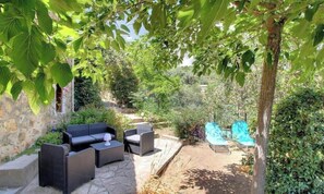 Un patio al aire libre con mobiliario de mimbre, un muro de piedra y un jardín con diversas plantas y una silla azul.