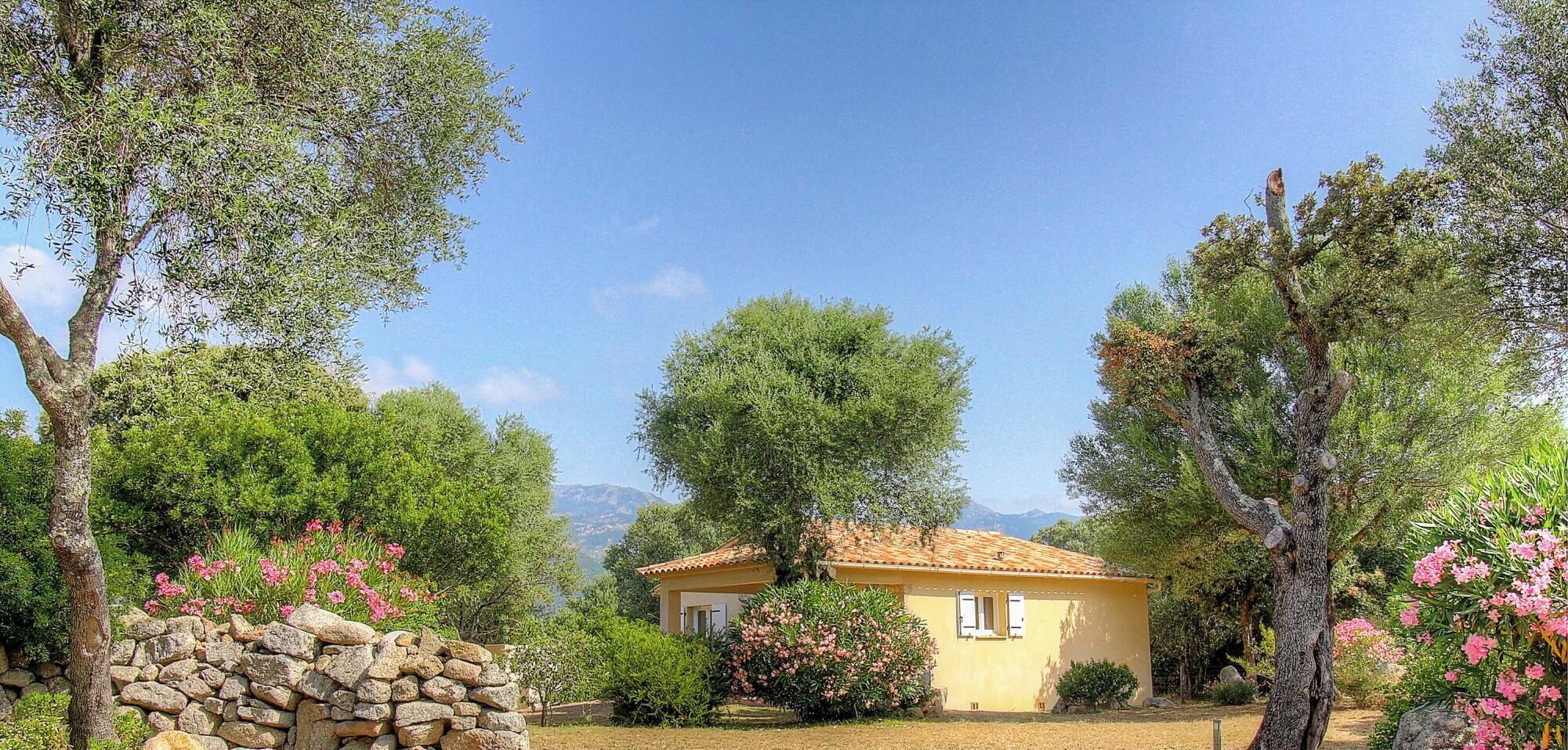 A yellow villa with a tiled roof, surrounded by trees and flowering plants.