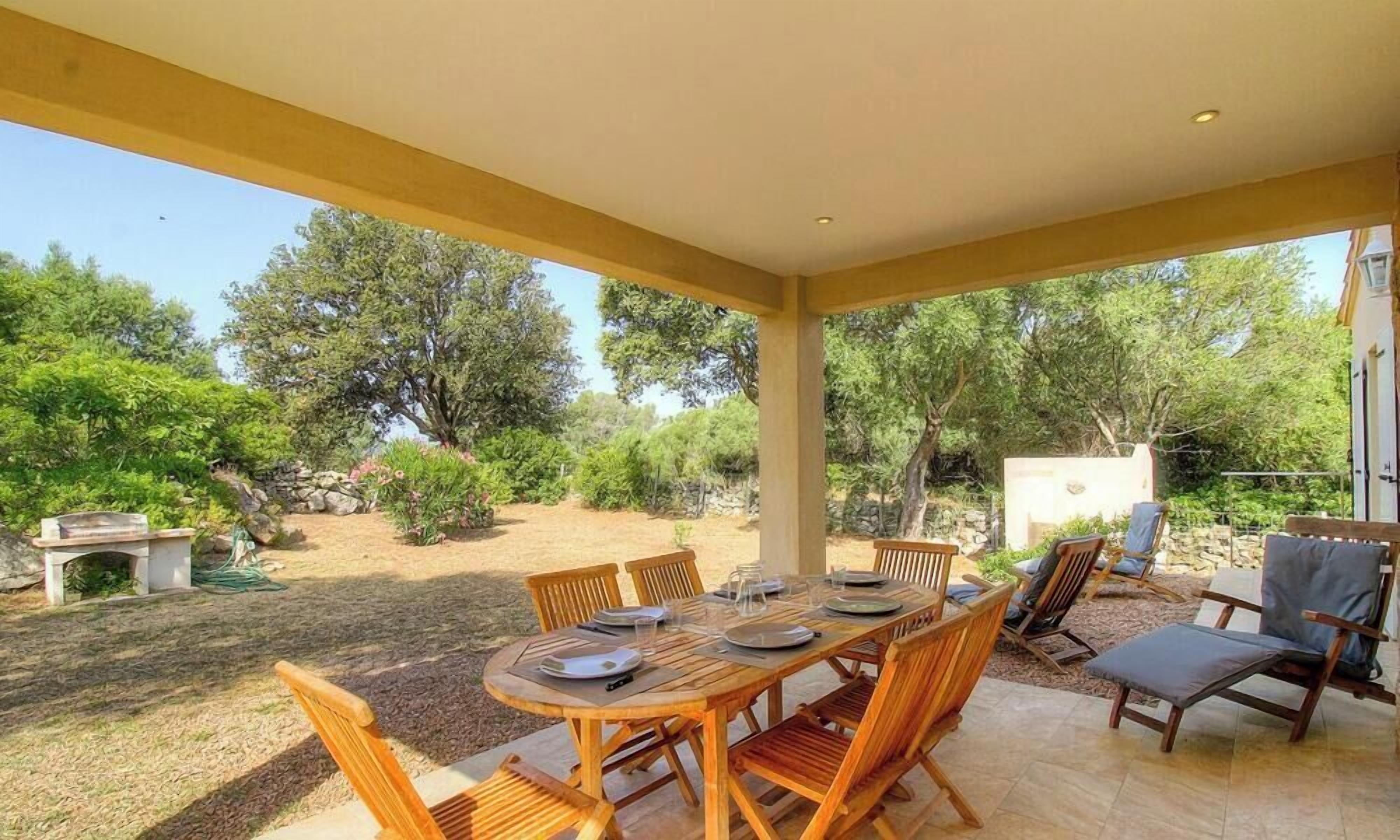 An outdoor patio with a wooden table and chairs, surrounded by greenery and a stone wall.