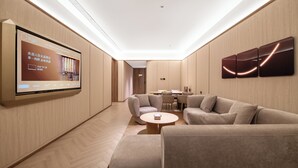 Una habitación de hotel moderna con un televisor de pantalla plana grande montado en la pared, un conjunto de sofás, una mesa redonda de centro y una zona de comedor con mesa y sillas.