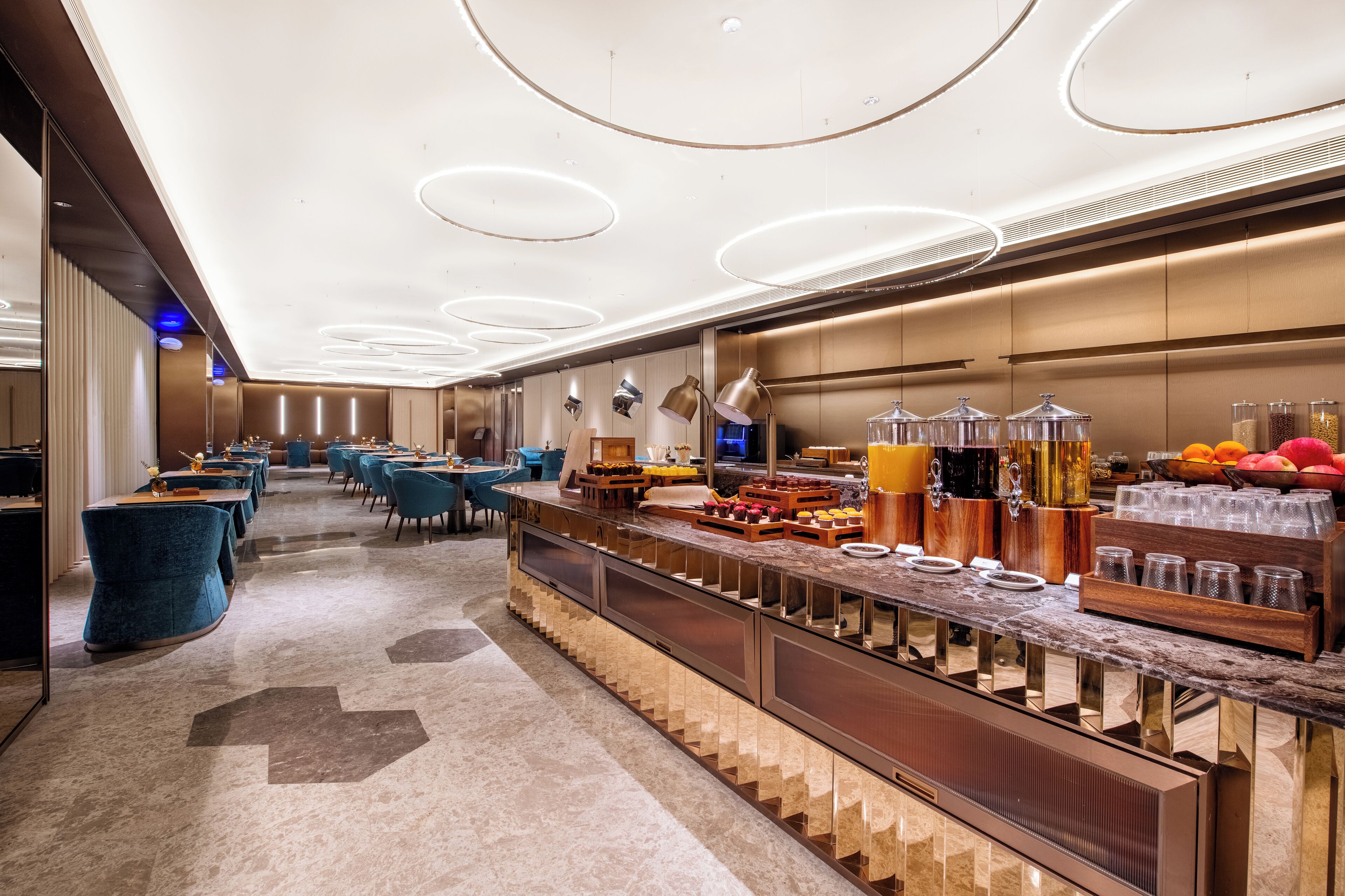 A modern hotel lobby with a long reception desk, blue chairs, and a buffet area with food and drinks.
