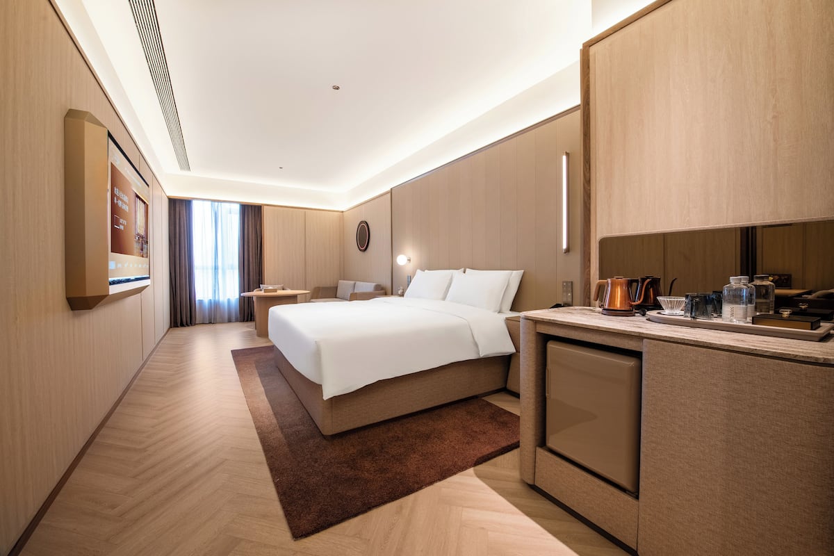 A modern hotel room with a large bed, a small kitchenette, a desk, and a view of the outdoors.
