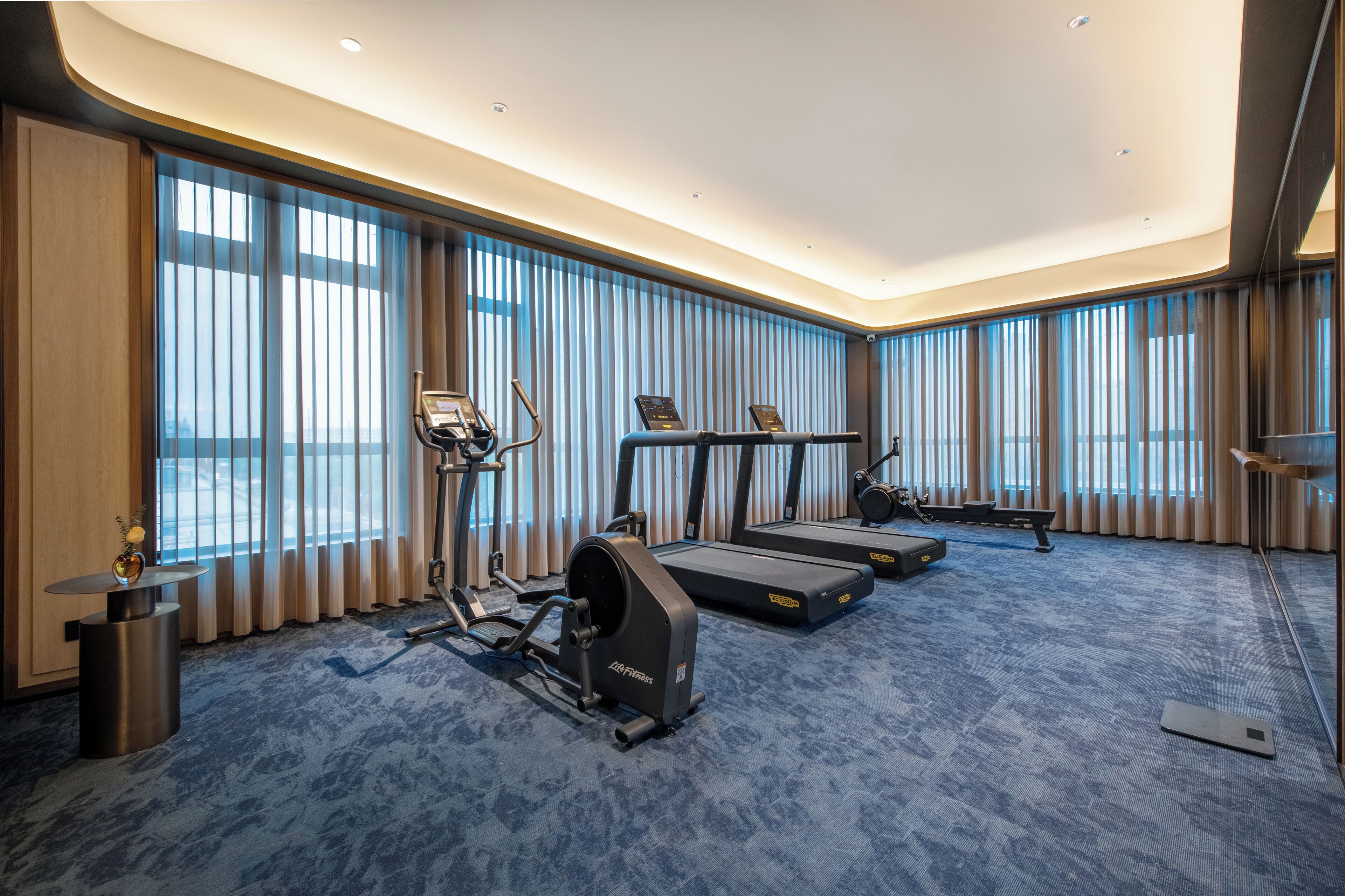 A hotel room with a large window, a treadmill, an elliptical machine, and a small table with a vase.